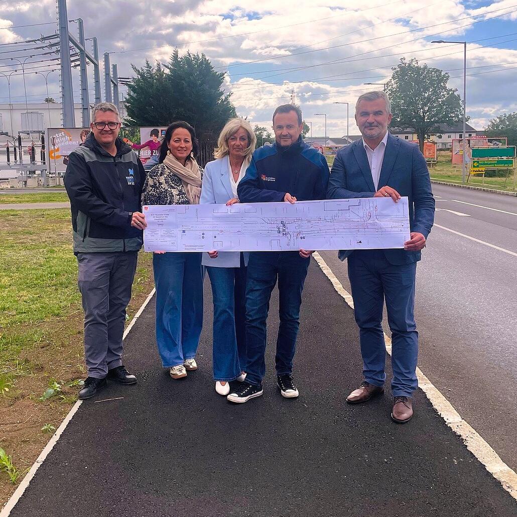 Fünf Personen halten einen Bauplan vor sich, im Hintergrund eine Straße und Stromleitungen.