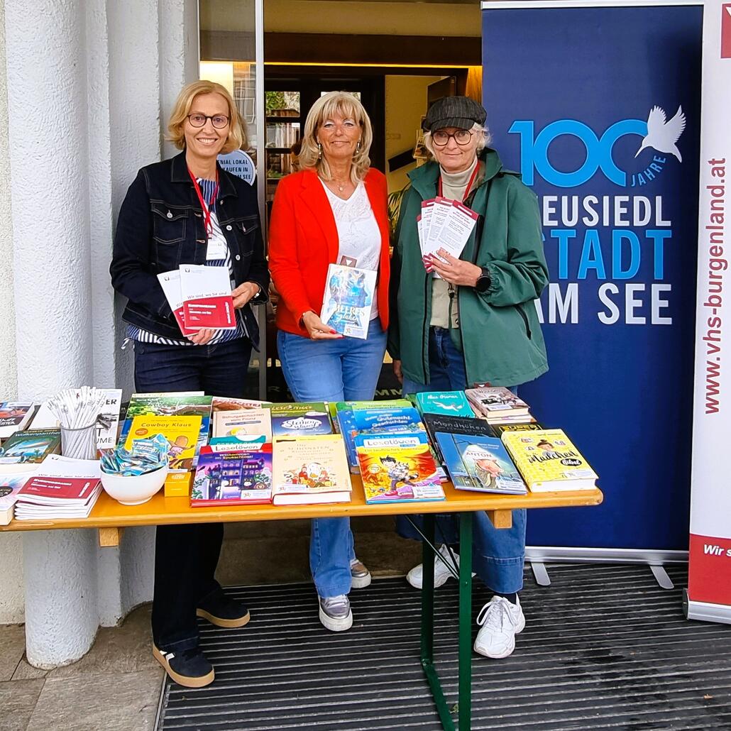 Drei Frauen präsentieren Bücher in Neusiedl am See vor einem Banner mit der Aufschrift "100 Jahre".